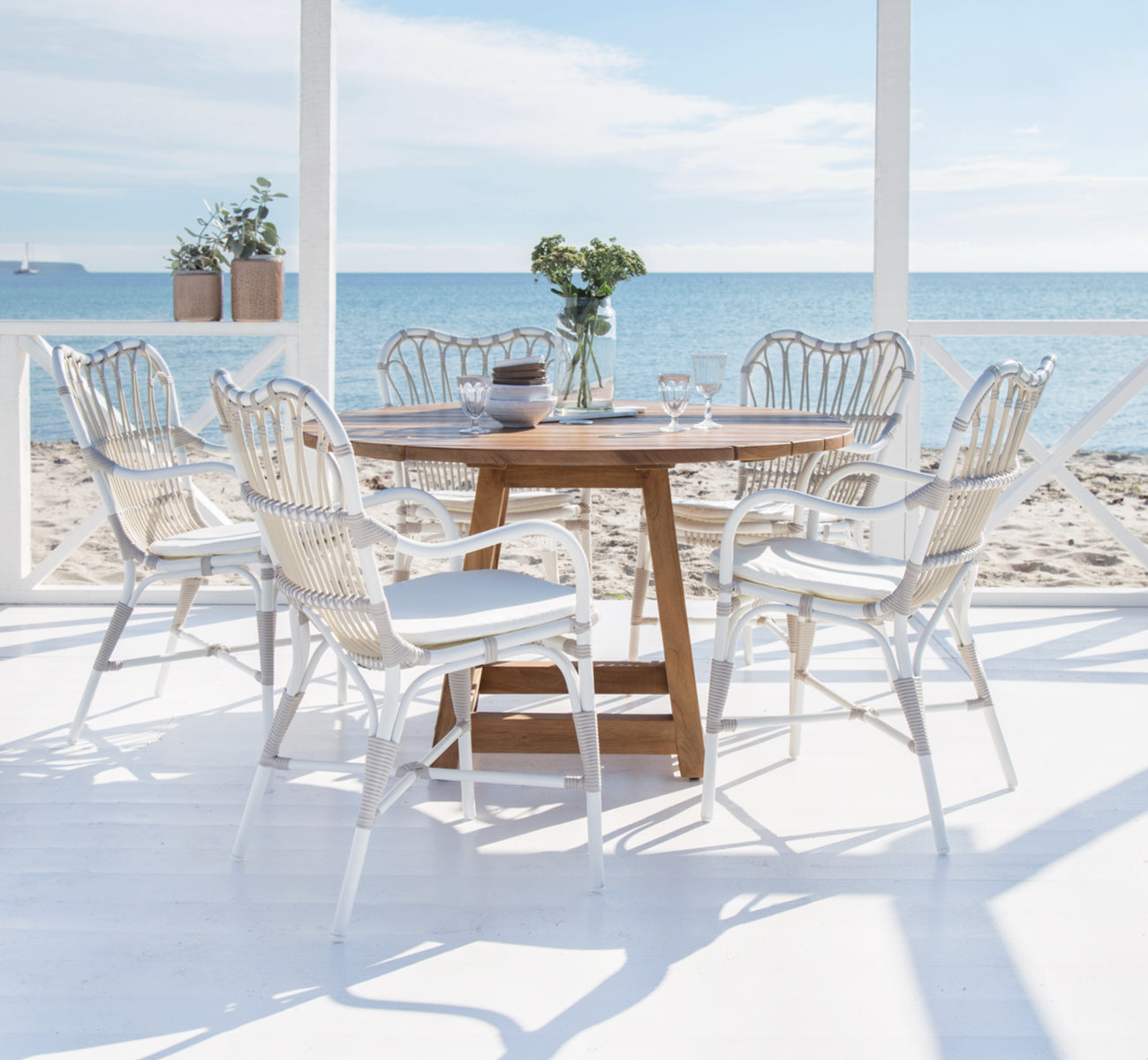 Dies ist ein Lifestyle-Bild von George Außentisch Ø160 in outdoor Teak, auf einer Gartenterrasse im Freien mit weißer Hausfassade und Bepflanzung.