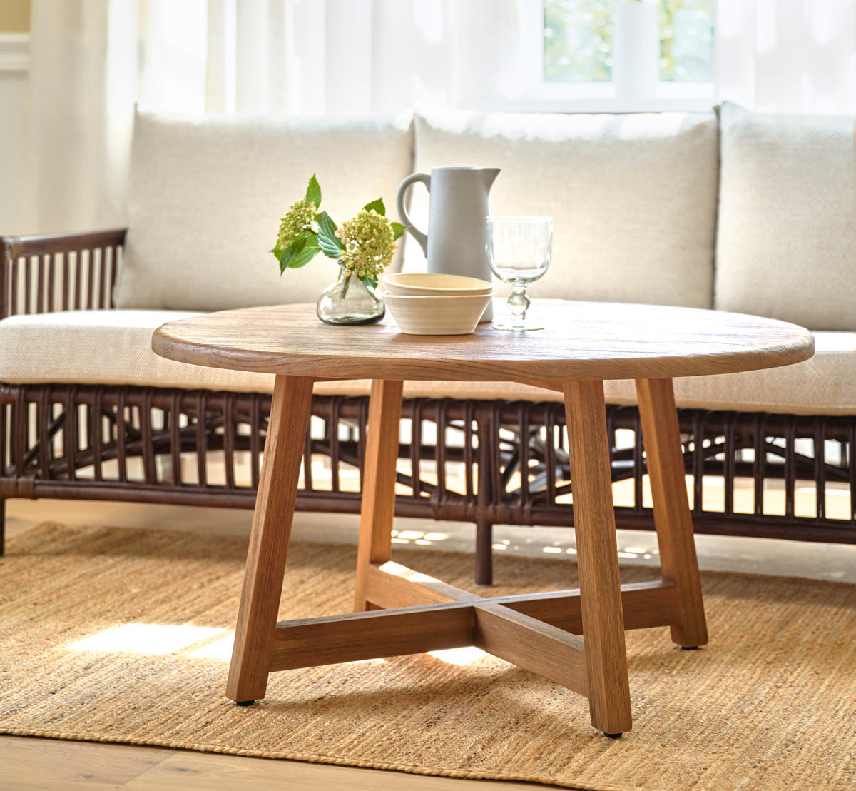 Dies ist ein Lifestyle-Bild von Aksel Couchtisch Ø100 cm in teak indoor. Vor einem Rattansofa auf einem Juteteppich.