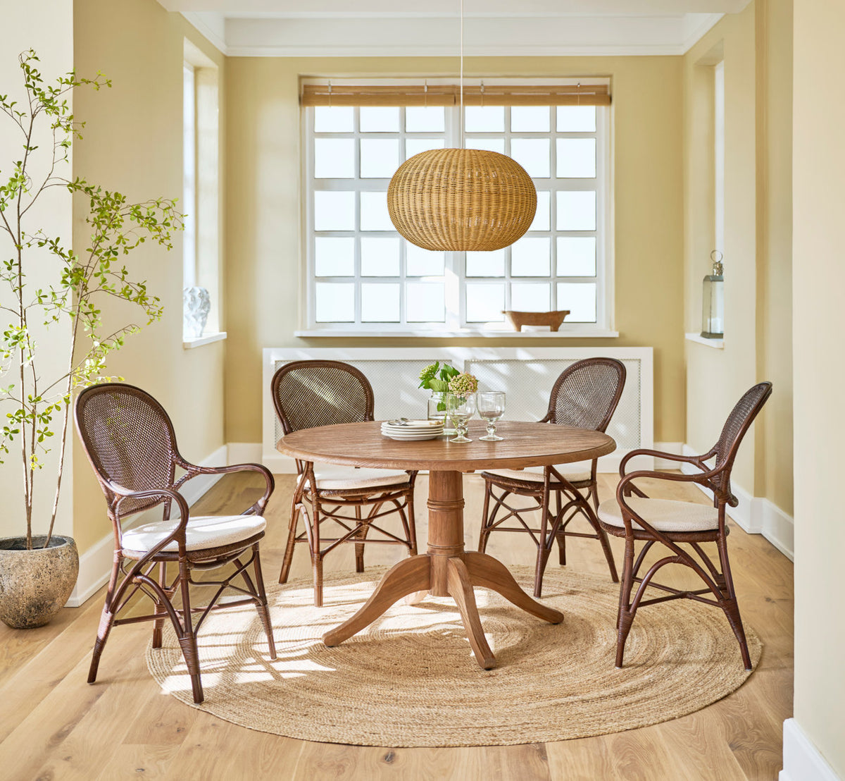 Dies ist ein Lifestyle-Bild von Michel Teak-Esstisch Ø120 cm in teak indoor. Mit Rattanstühlen und Pendelleuchte im sonnigen Esszimmer.