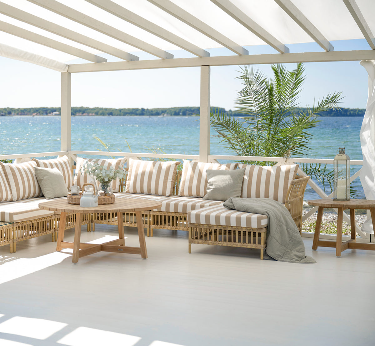 Dies ist ein Lifestyle-Bild von Victor Teak Lounge Table Ø75 cm in teak outdoor. Auf einer Strandterrasse zwischen Tagesbetten mit gestreiften Kissen aufgestellt.
