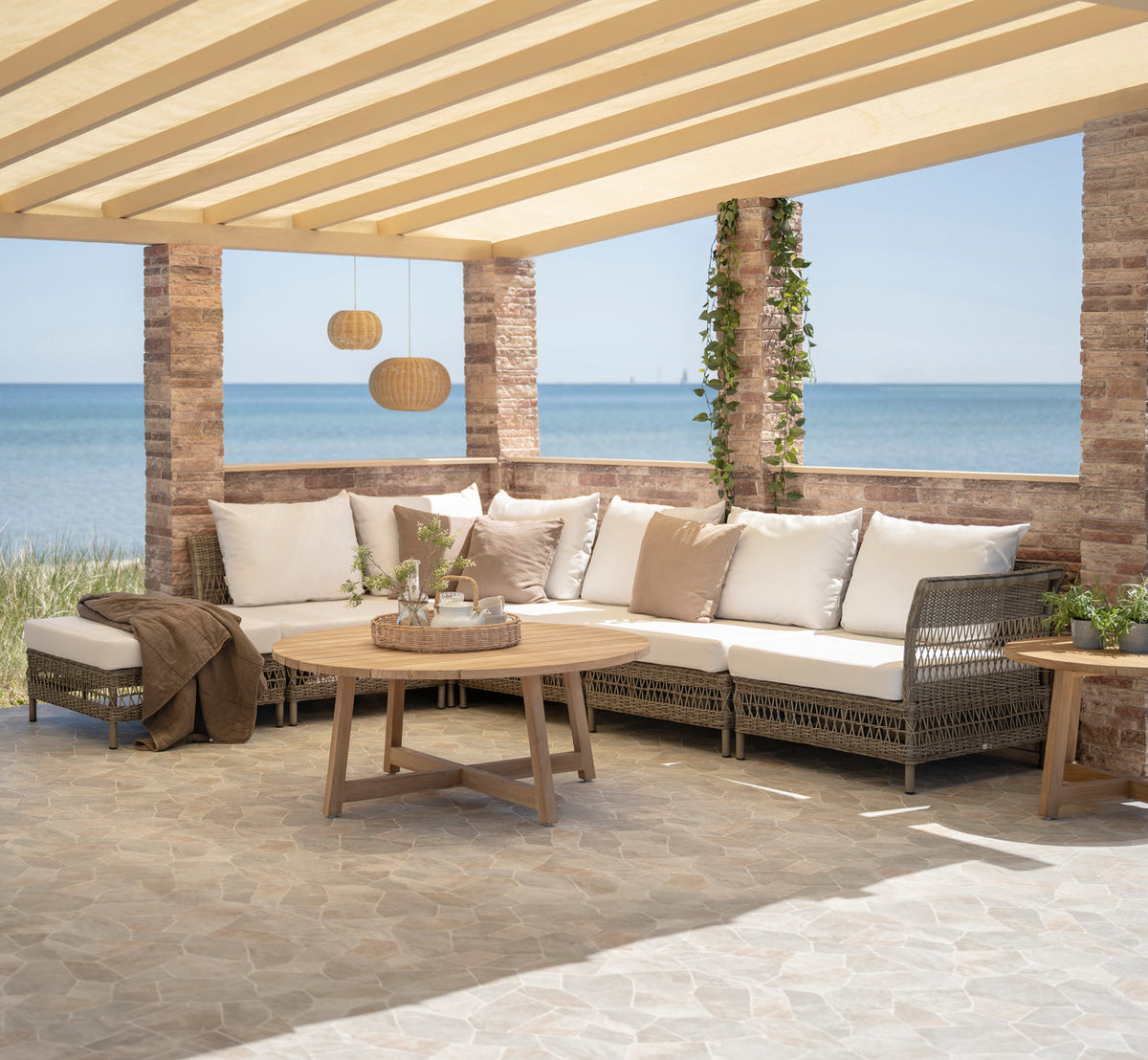 Dies ist ein Lifestyle-Bild von Victor Teak-Loungetisch Ø75 cm in teak outdoor. Ecksofa, Pouf und Teaktisch unter einer Pergola am Meer.