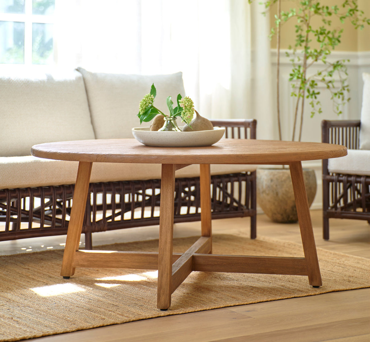 Dies ist ein Lifestyle-Bild von Aksel Couchtisch Ø120 cm in teak indoor. Vor einem Rattansofa in einem sonnigen Wohnzimmer.