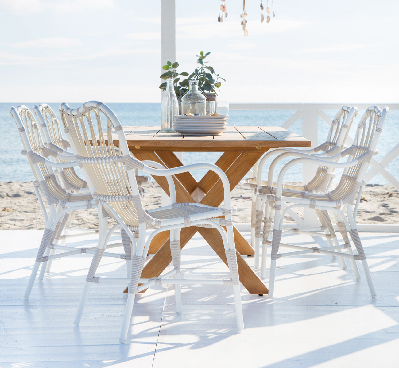 Dies ist ein Lifestyle-Bild von Colonial Tisch 160 in outdoor Teak, auf einer Strandterrasse mit weißem Holzdeck und Meerblick.