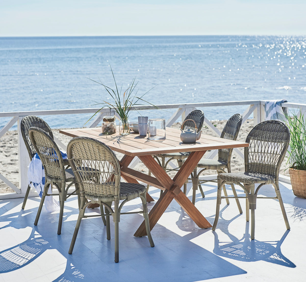 Dies ist ein Lifestyle-Bild von Colonial Tisch 160 in outdoor Teak, auf einer Strandterrasse mit weißem Holzdeck und Meerblick.