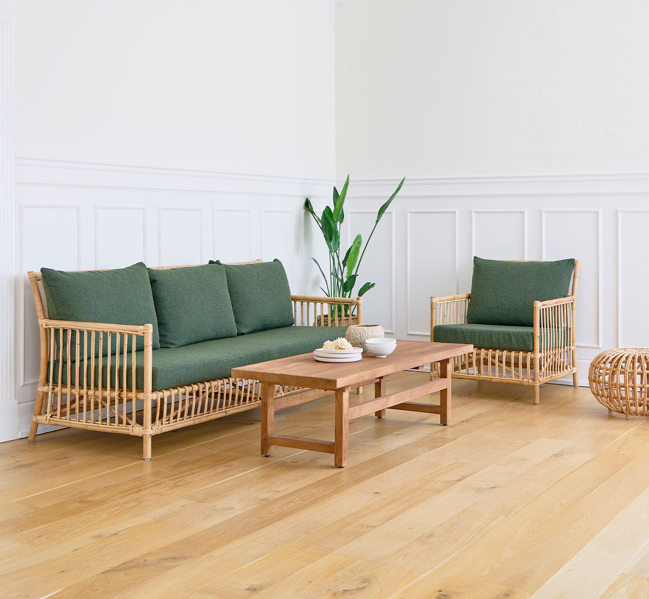 Dies ist ein Lifestyle-Bild von Alfred Couchtisch in teak indoor, in einem hellen Wohnzimmer mit weißen Wandpaneelen und einem Sofa mit grünen Kissen.