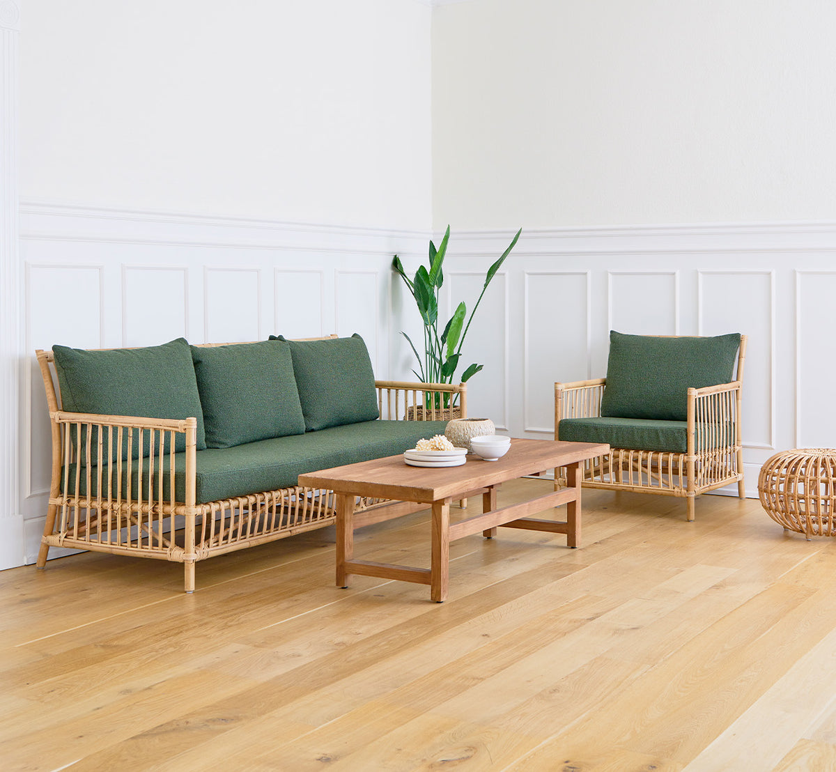 Dies ist ein Lifestyle-Bild von Alfred Couchtisch in teak indoor, in einem hellen Wohnzimmer mit weißen Wandpaneelen und einem Sofa mit grünen Kissen.
