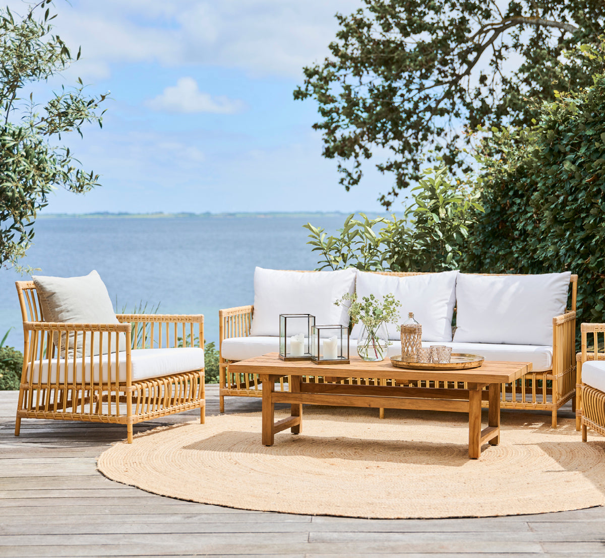 Dies ist ein Lifestyle-Bild von Julian Teak Lounge Table 140x55 cm in teak outdoor. Auf einer Terrasse mit Loungemöbeln, einem Juteteppich und Meerblick.