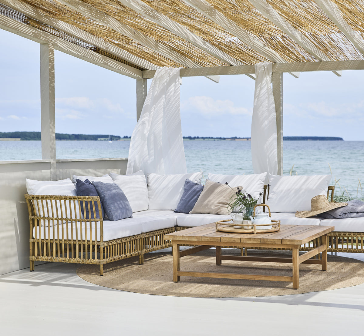 Dies ist ein Lifestyle-Bild von Julian Teak Lounge Table 120x120 cm in teak outdoor. Unter einer Strandpergola mit Loungesitzplätzen und Meerblick.