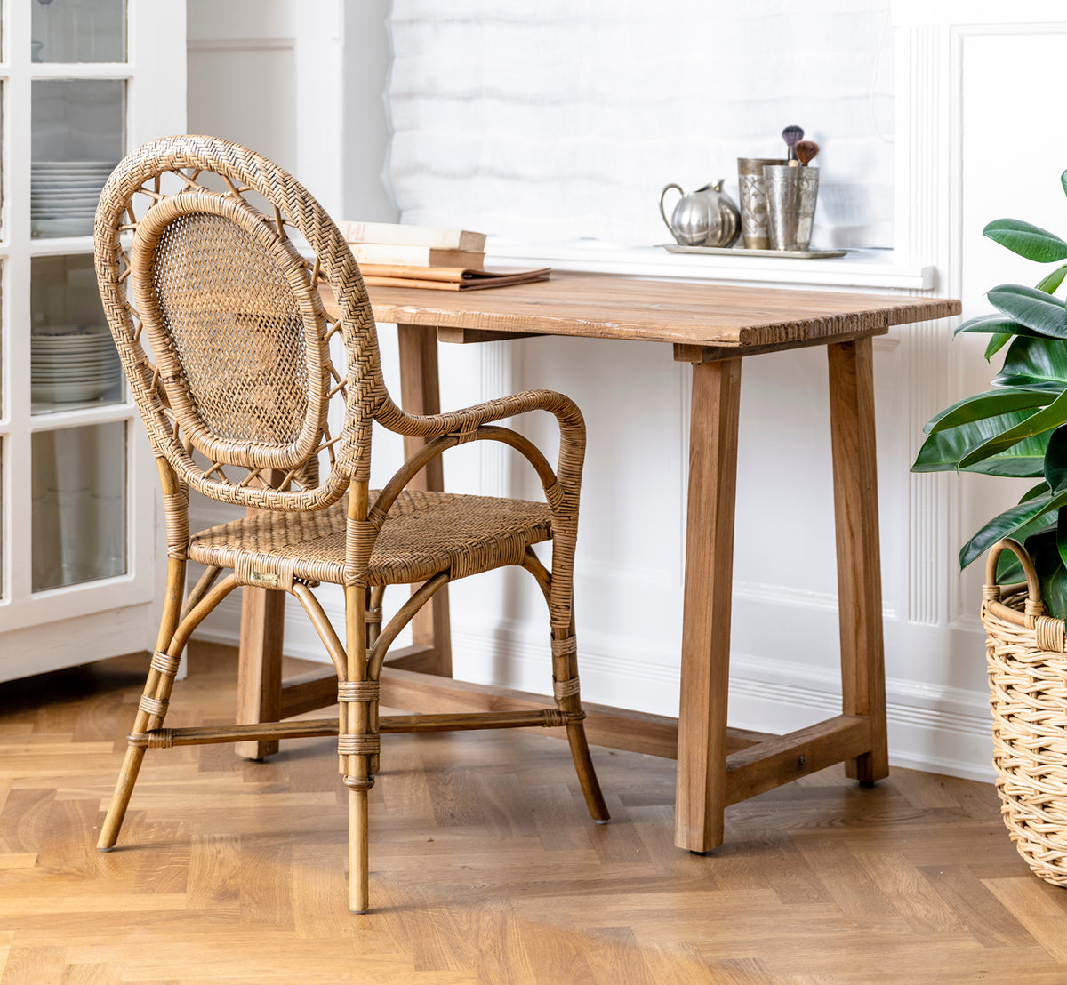 Dies ist ein Lifestyle-Bild von Lucas Teak Schreibtisch in teak indoor. Mit einem Rattanstuhl und grüner Pflanze in einem hellen Interieur mit Fischgrätboden kombiniert.