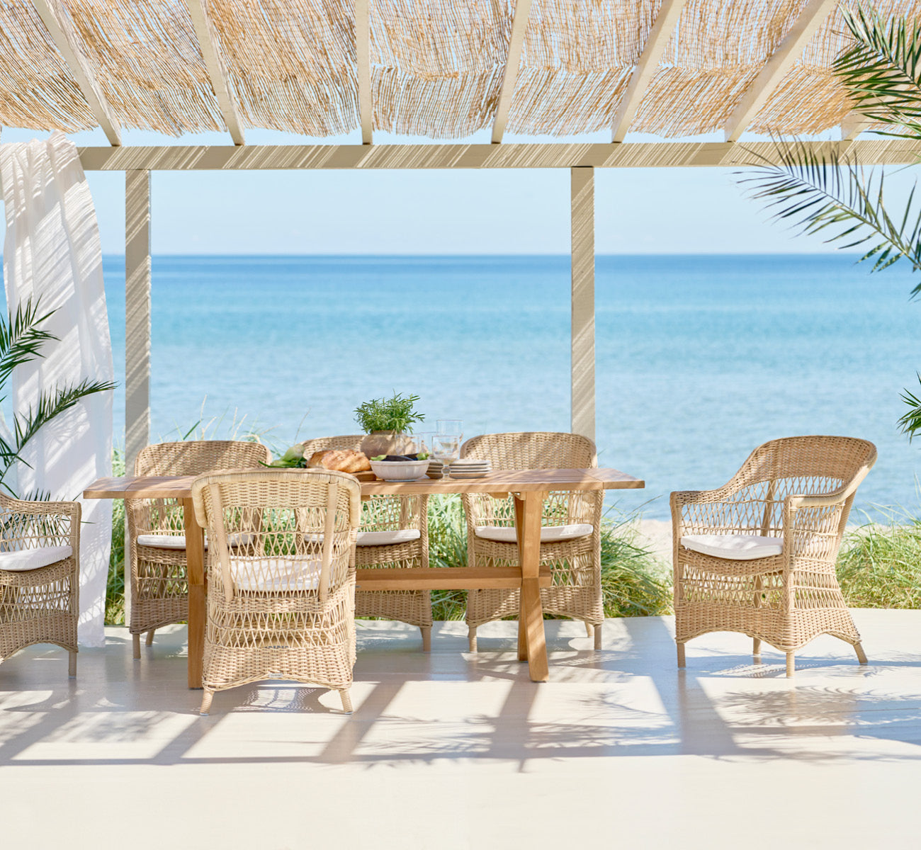 Dies ist ein Lifestyle-Bild von Colonial Tisch 200 in outdoor Teak, auf einer Strandterrasse mit weißem Holzdeck und Meerblick.