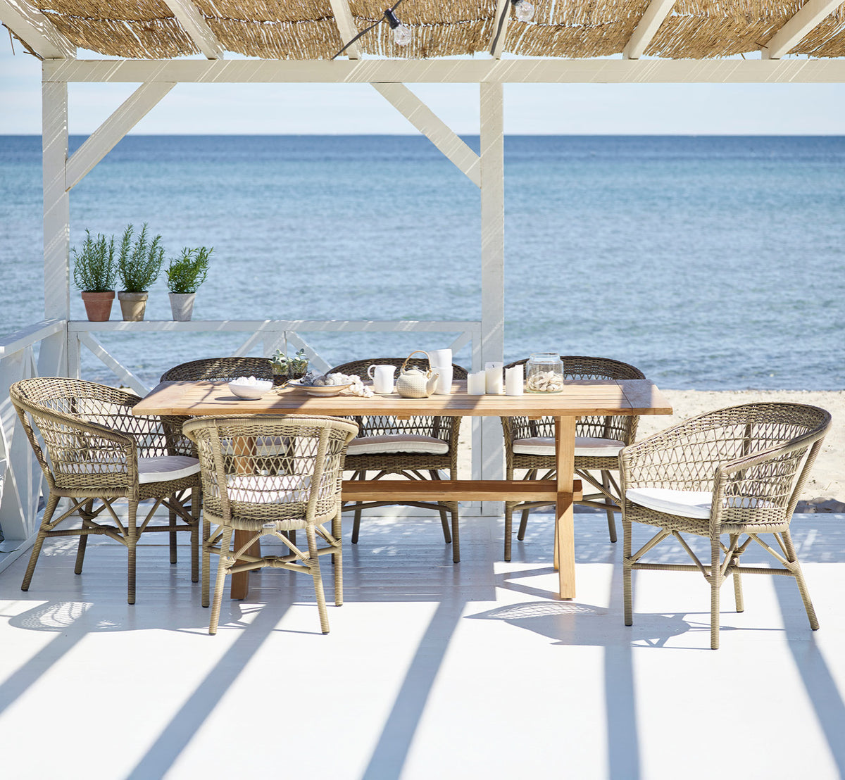 Dies ist ein Lifestyle-Bild von Colonial Tisch 200 in outdoor Teak, auf einer Strandterrasse mit weißem Holzdeck und Meerblick.