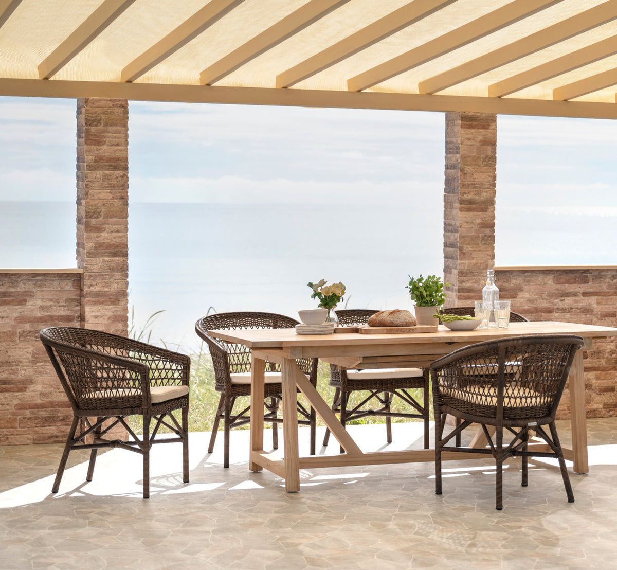 Dies ist ein Lifestyle-Bild von George Teak-Ausziehtisch 200-280x100 cm in teak outdoor. Zum Mittagessen gedeckt auf einer Terrasse am Meer.