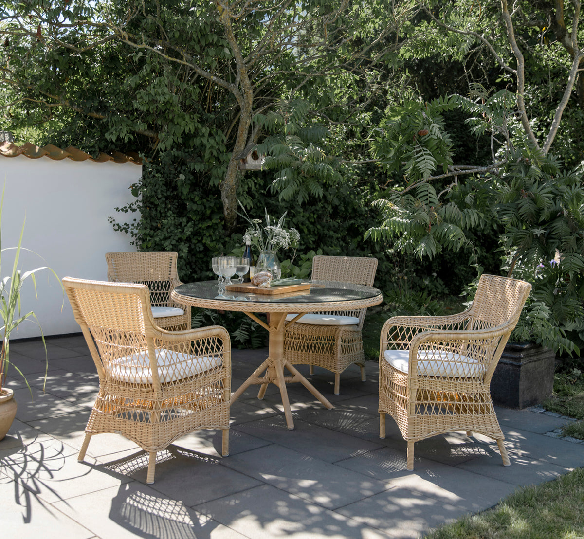 Dies ist ein Lifestyle-Bild von Grace Außen-Esstisch in natural ArtFibre Aluminium, auf einer üppigen Gartenterrasse mit Steinboden und überhängenden.