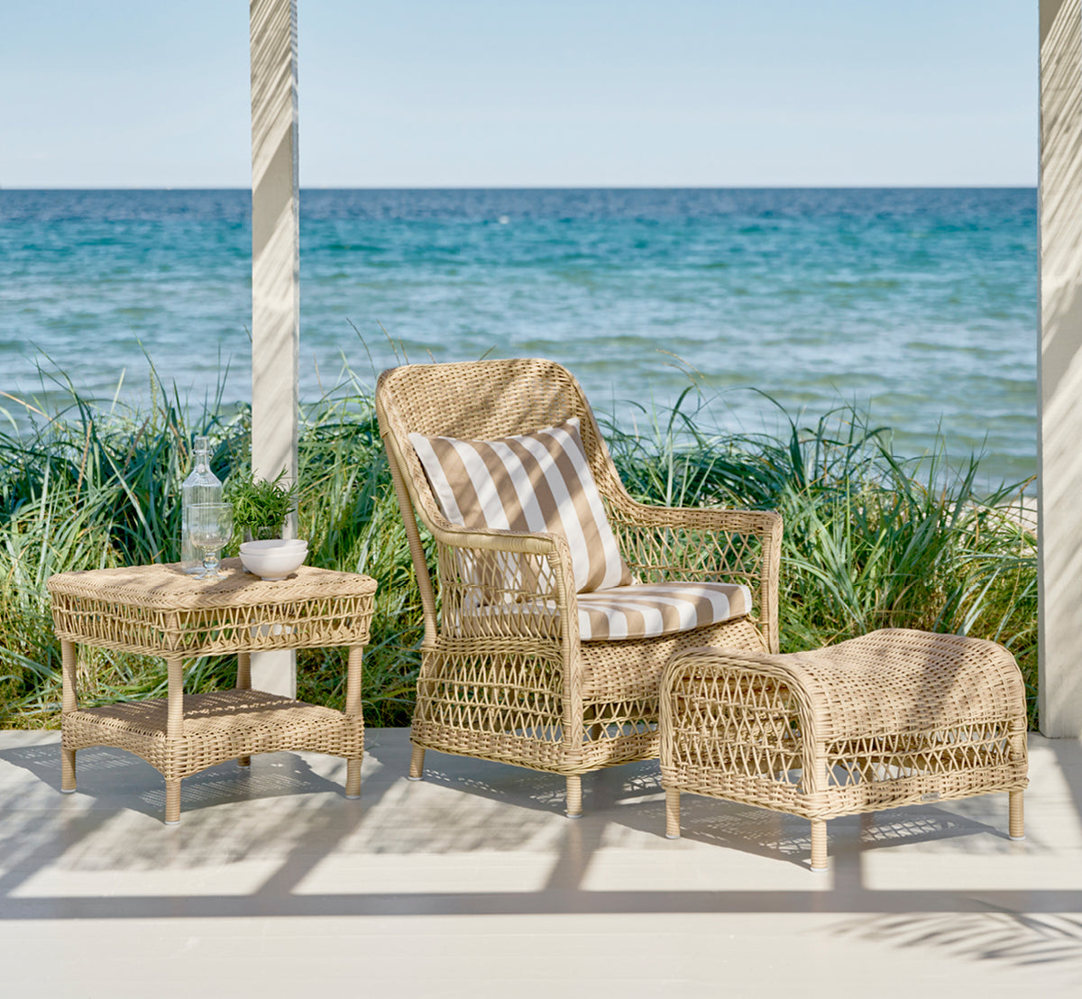 Dies ist ein Lifestyle-Bild von Susy Beistelltisch in natural ArtFibre Aluminium, auf einer Küstenterrasse mit Meerblick, Strandgräsern und weißen.