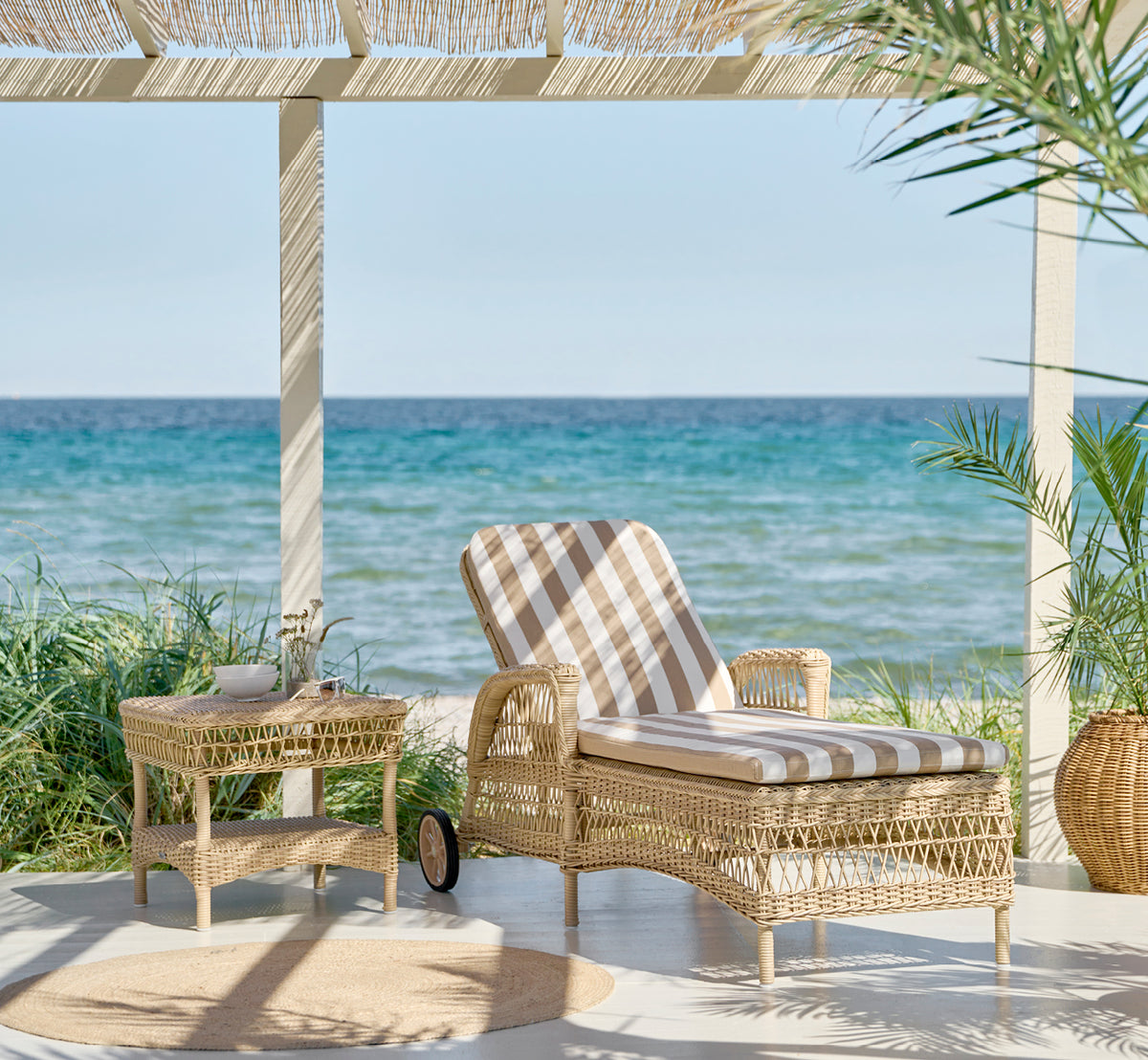 Dies ist ein Lifestyle-Bild von Susy Beistelltisch in natural ArtFibre Aluminium, auf einer Küstenterrasse mit Meerblick, Strandgräsern und weißen.