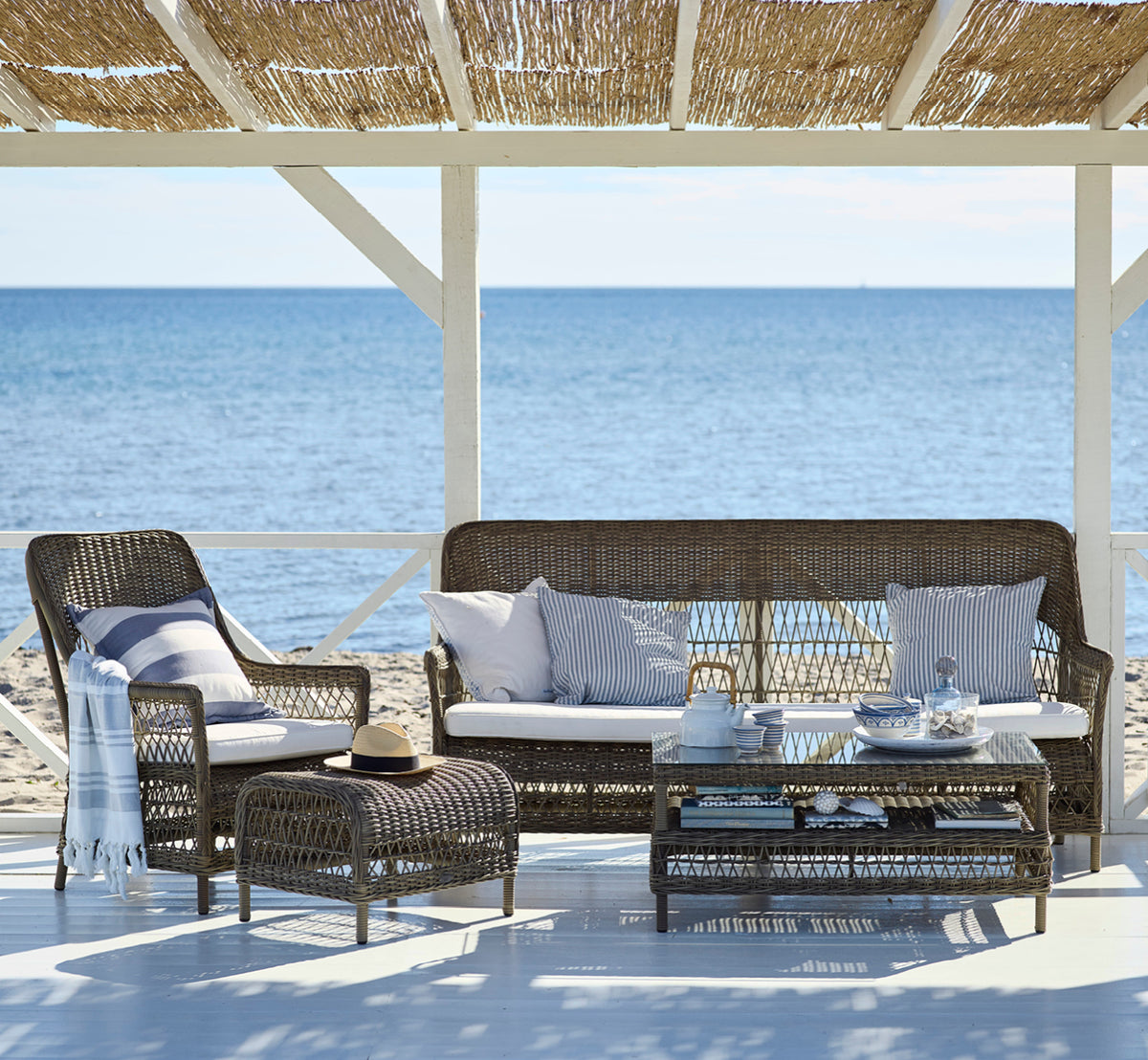 Dies ist ein Lifestyle-Bild von Hazel Couchtisch in antique Grey ArtFibre Aluminium, auf einer schattigen Küstenterrasse mit Schilfdach und Meerblick.