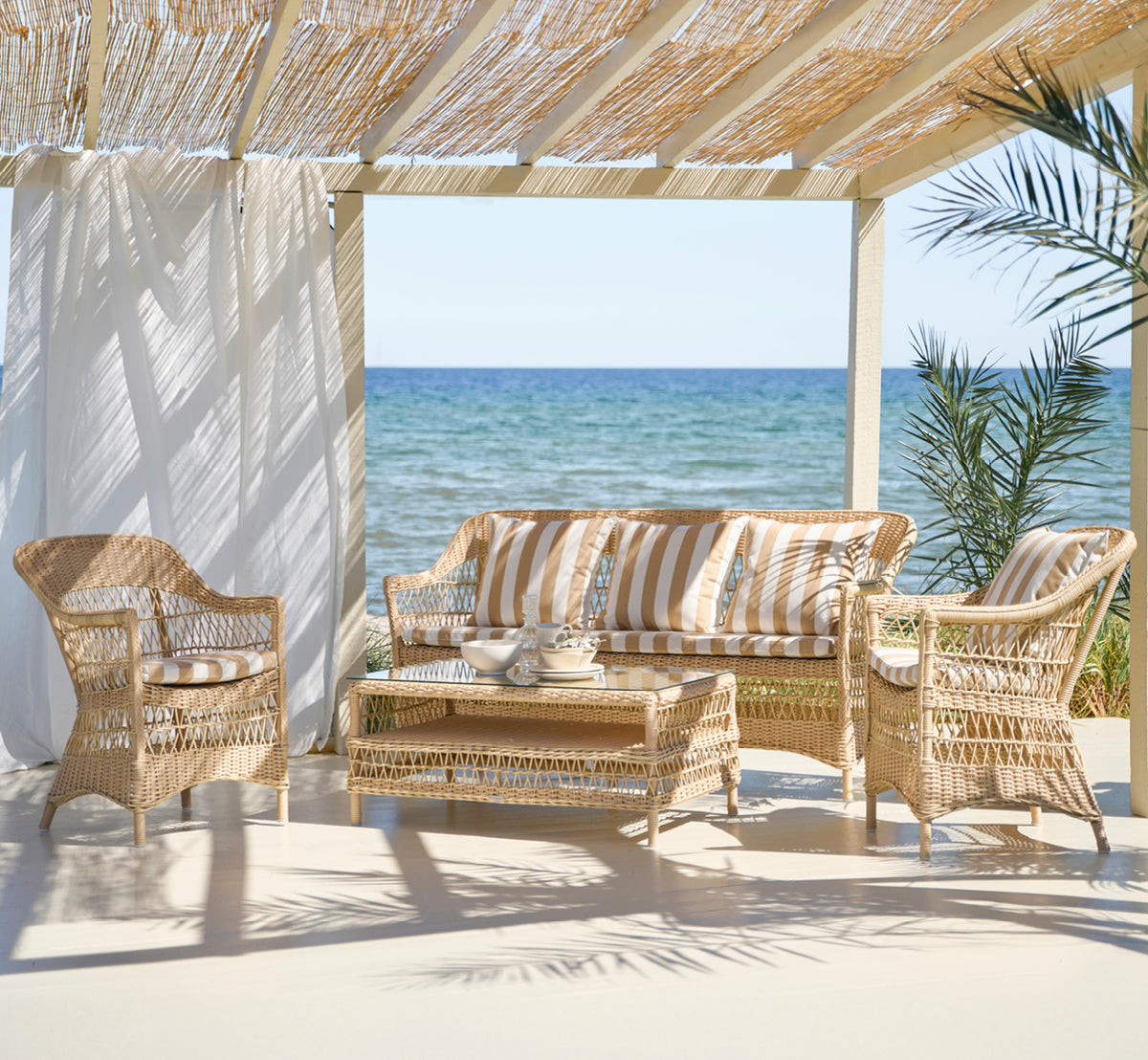 Dies ist ein Lifestyle-Bild von Hazel Couchtisch in natural ArtFibre Aluminium, auf einer schattigen Küstenterrasse mit Schilfdach und Meerblick.