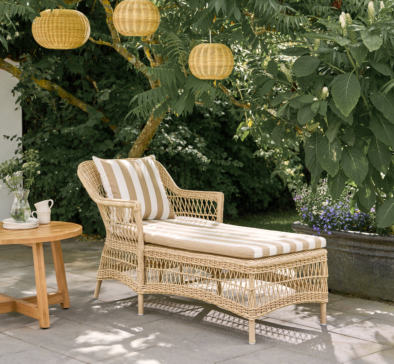 Dies ist ein Lifestyle-Bild von Olivia Exterior Sunbed i natural artfibre aluminium. Auf einer Terrasse mit gestreiftem Kissen und Laternen.