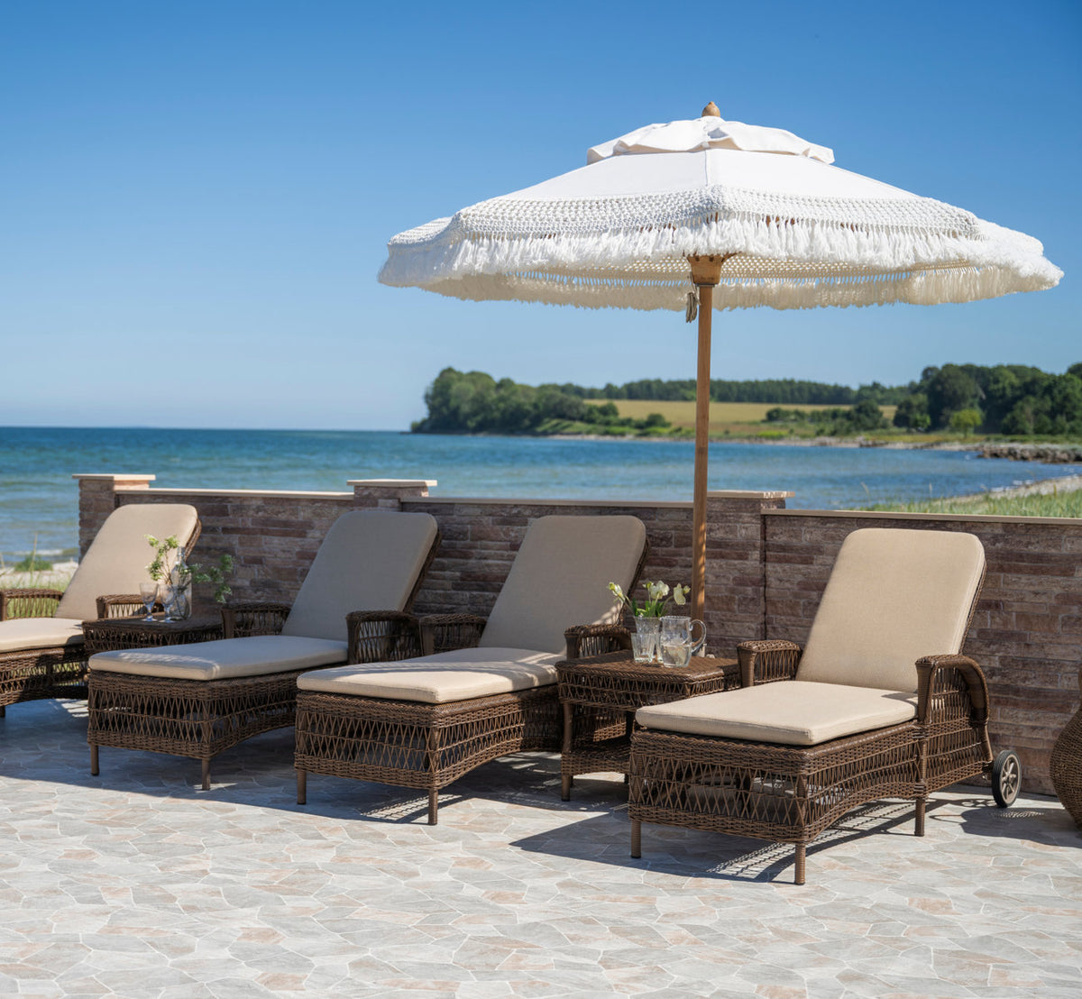 Dies ist ein Lifestyle-Bild von Daisy Außen-Sonnenliege in brown artfibre aluminum. Reihe von Liegen unter einem Fransenschirm am Meer.