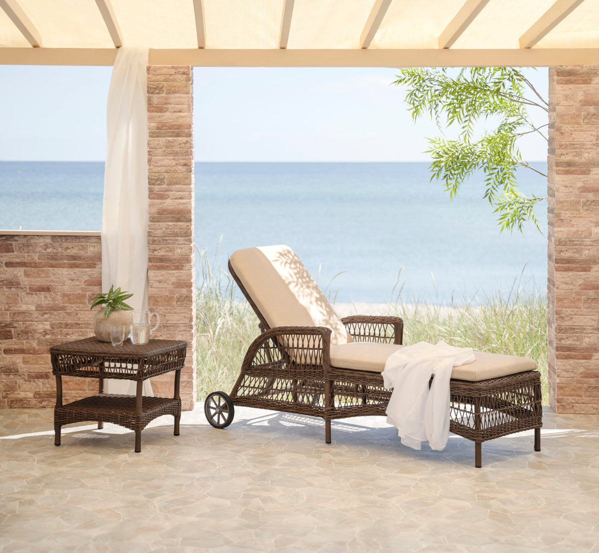 Dies ist ein Lifestyle-Bild von Daisy Außen-Sonnenliege in brown artfibre aluminum. Mit weißem Handtuch auf einer Terrasse am Meer.
