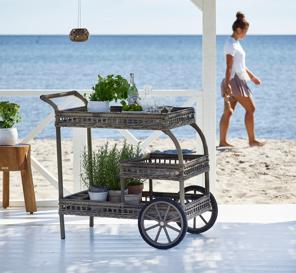 Dies ist ein Lifestyle-Bild von James Servierwagen in antique grey artfibre aluminium, auf einer Strandterrasse unter einer weißen Pergola mit.