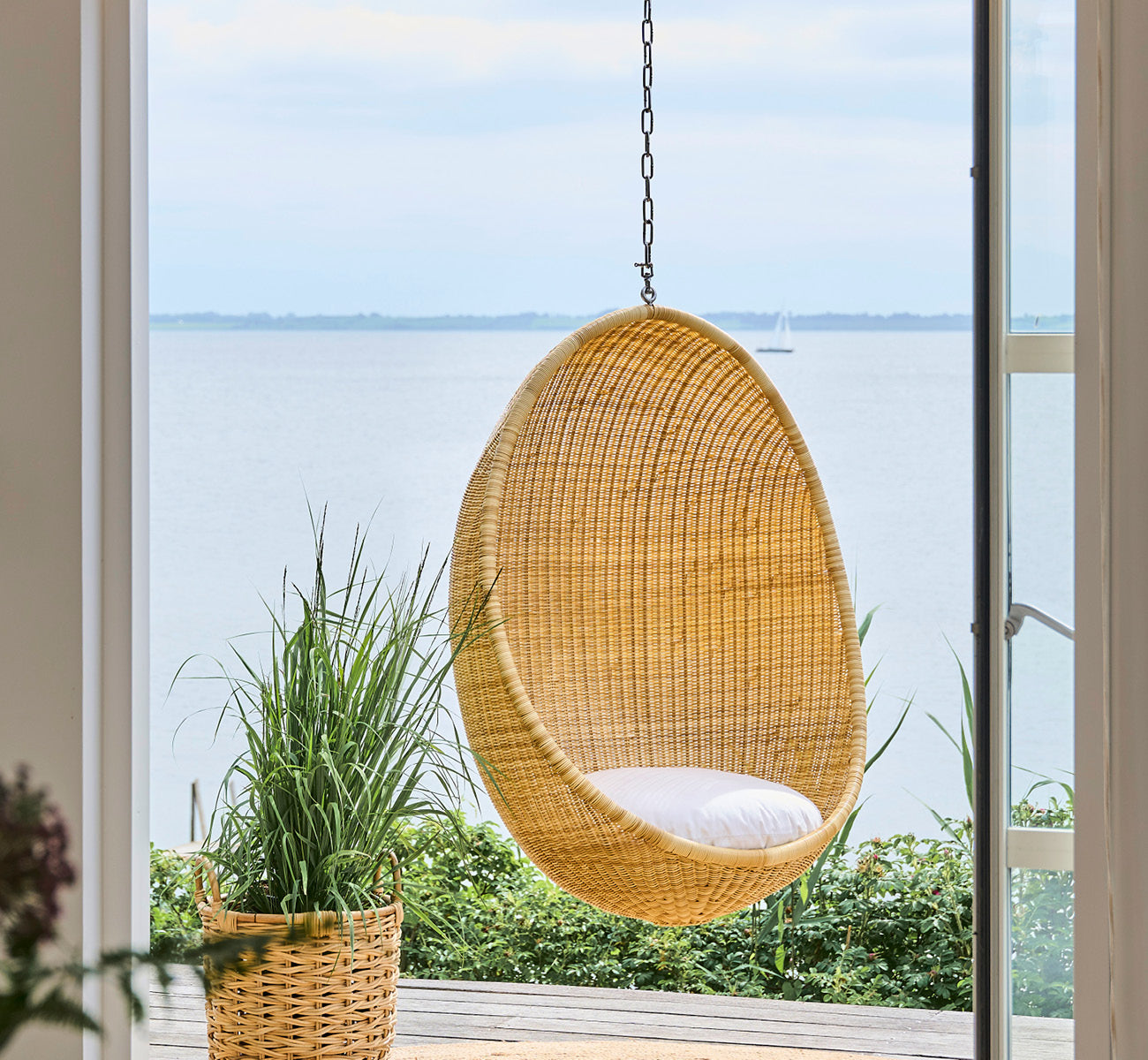 Dies ist ein Lifestyle-Bild von Chain Hänge-Ei in metal, hängend an einer Glastür mit Wasserblick und umgebenden grünen Pflanzen.