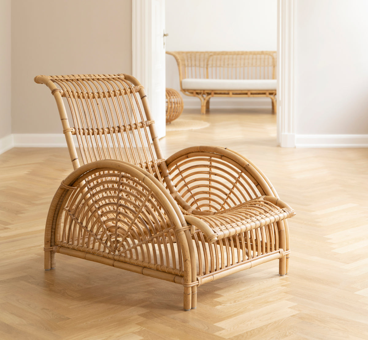 Dies ist ein Lifestyle-Bild von Paris Lounge-Sessel in polished natural rattan. Auf einem Fischgrätparkettboden neben einer passenden Bank platziert.