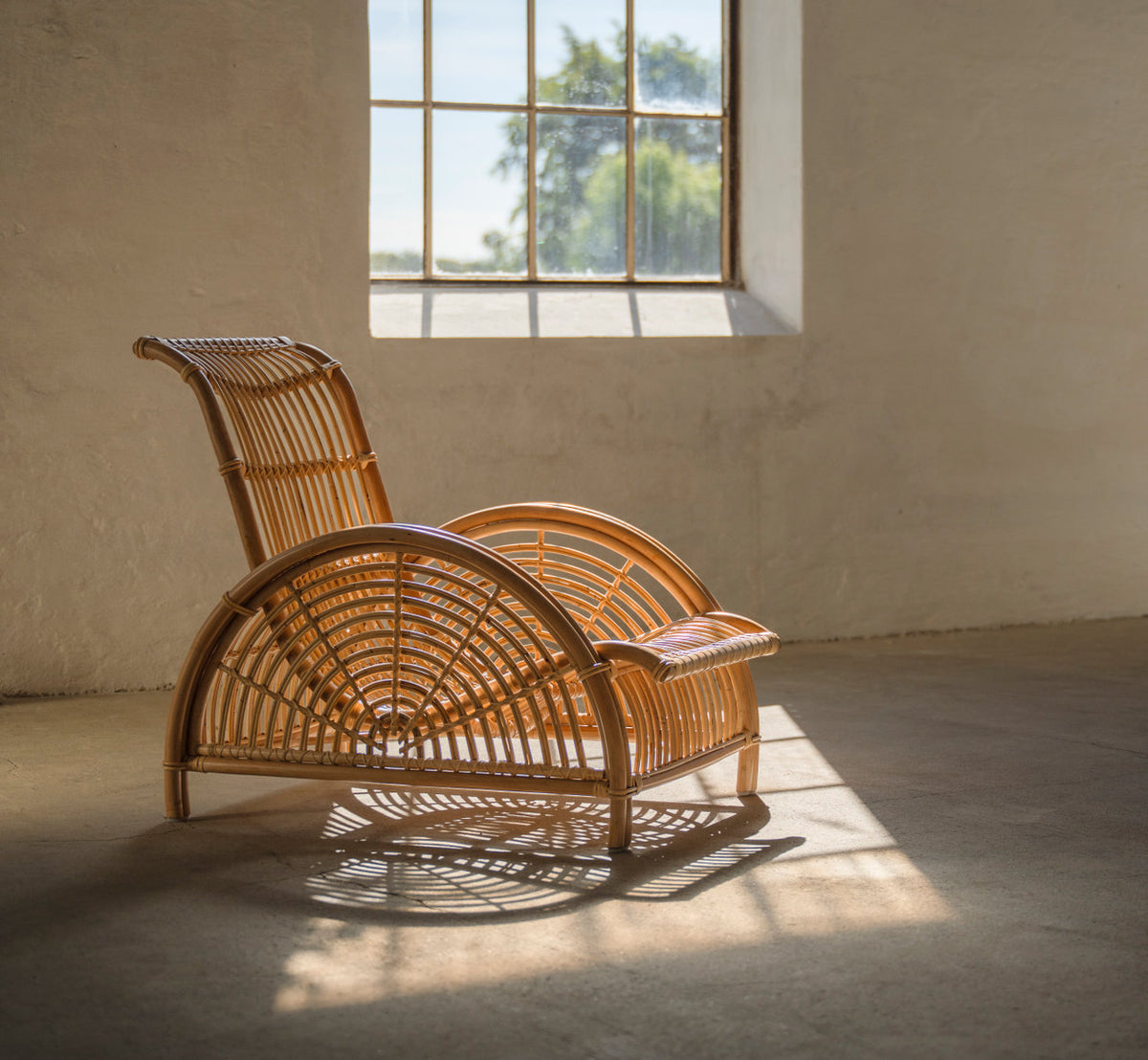 Dies ist ein Lifestyle-Bild von Paris Lounge-Sessel in polished natural rattan. Im warmen Sonnenlicht am Fenster in einem rustikalen Raum.