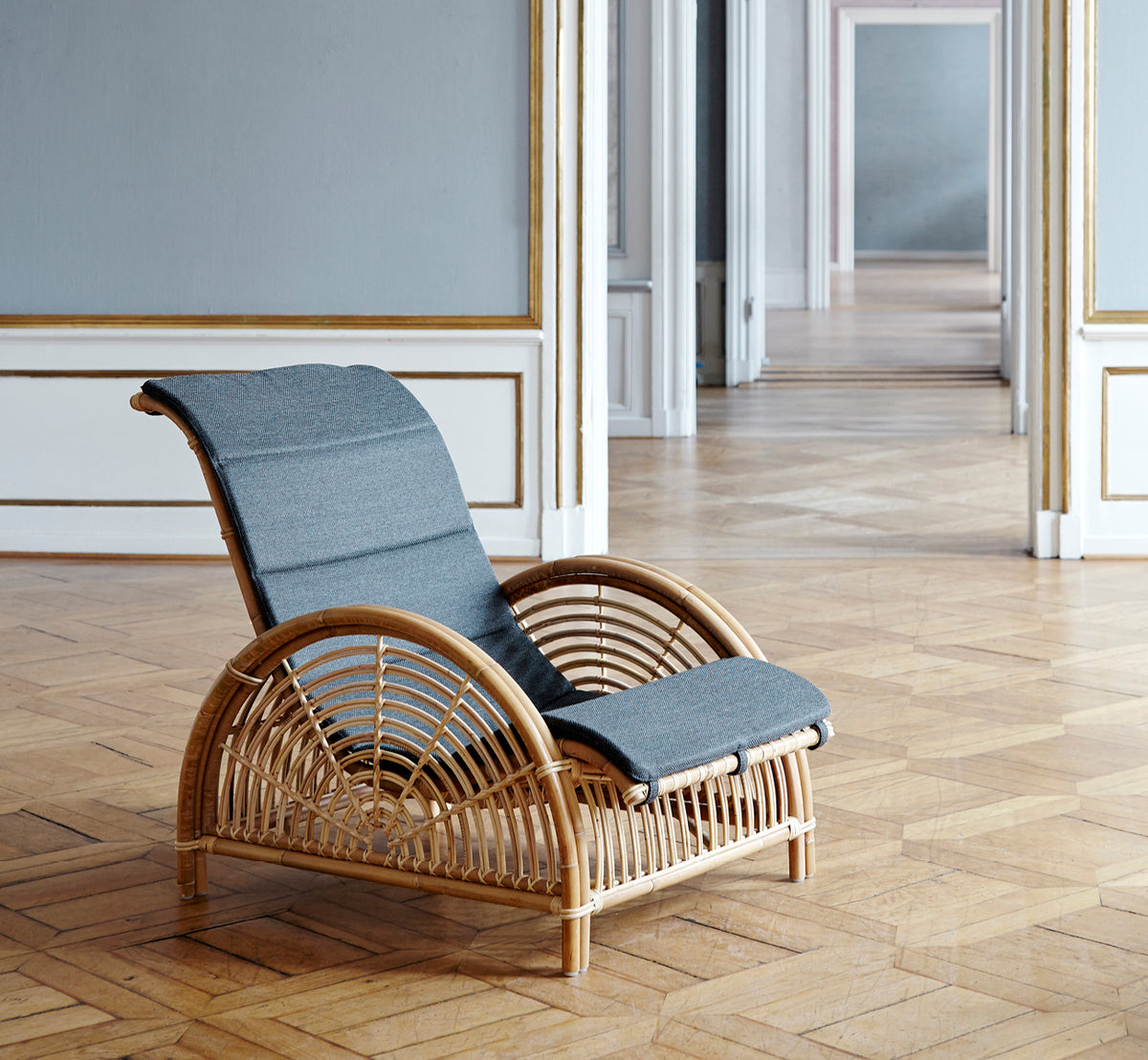 Dies ist ein Lifestyle-Bild von Paris Lounge-Sessel in polished natural rattan. Auf einem Fischgrätparkettboden neben einer passenden Bank platziert.