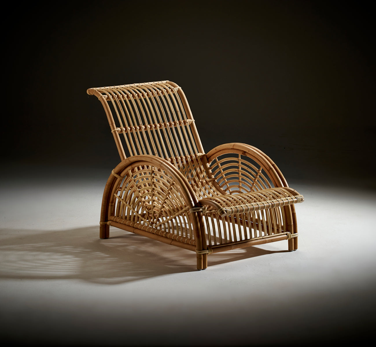 Dies ist ein Lifestyle-Bild von Paris Lounge-Sessel in polished natural rattan. Auf einem Fischgrätparkettboden neben einer passenden Bank platziert.