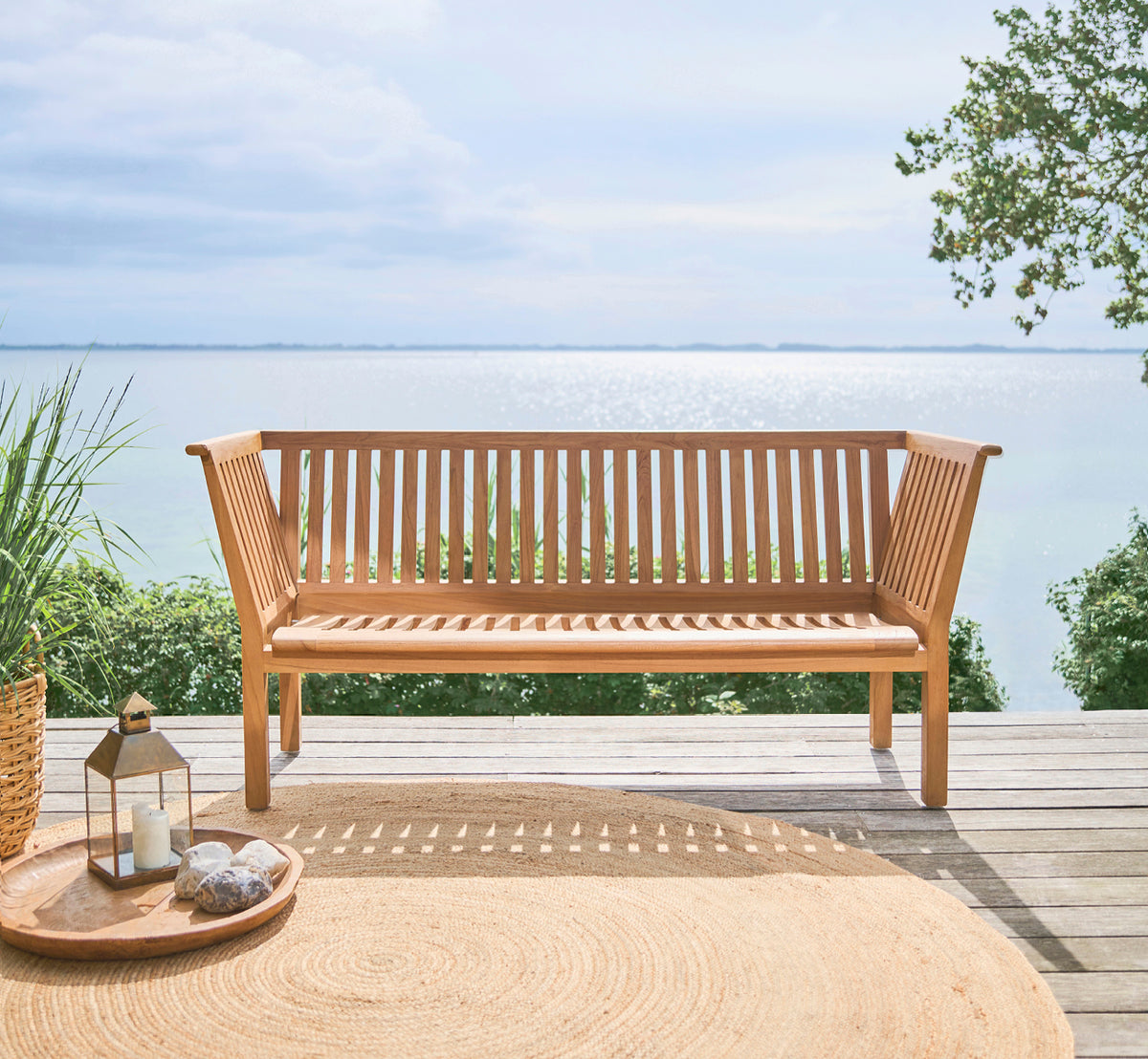 Dies ist ein Lifestyle-Bild von St Catherine Teakbank in teak outdoor. Auf einer Holzterrasse mit Meerblick und einem runden Juteteppich aufgestellt.
