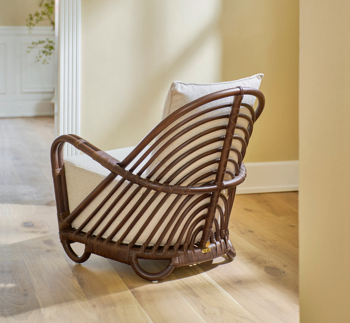 Dies ist ein Lifestyle-Bild von Charlottenborg Lounge-Sessel in brown rattan. Mit cremefarbenem Kissen auf Eichenboden an einer Tür.