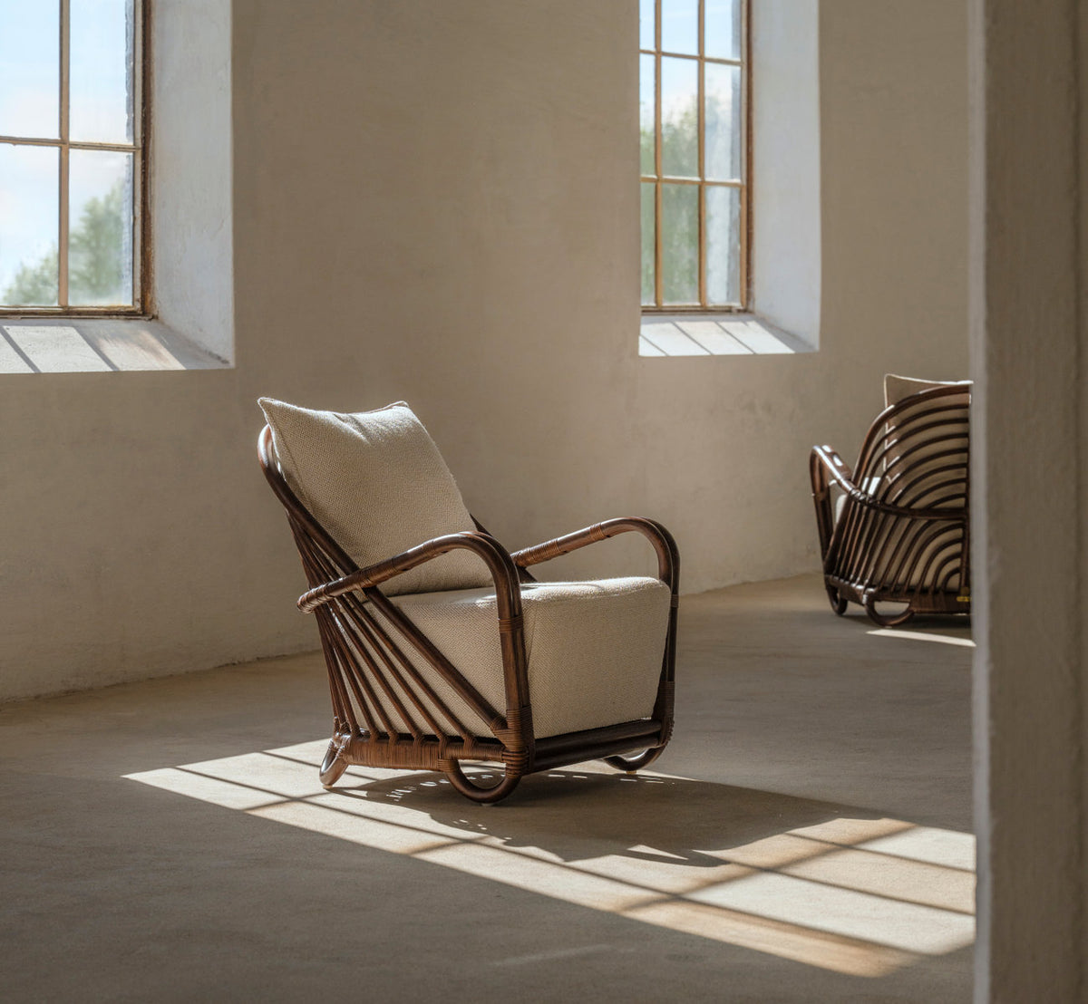 Dies ist ein Lifestyle-Bild von Charlottenborg Lounge-Sessel in brown rattan. Mit Kissen in einem sonnigen Raum mit Betonboden.
