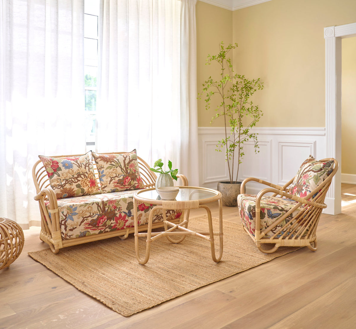 Dies ist ein Lifestyle-Bild von Charlottenborg Couchtisch in polished natural rattan. Mit geblümten Kissen in einem hellen Wohnzimmer.
