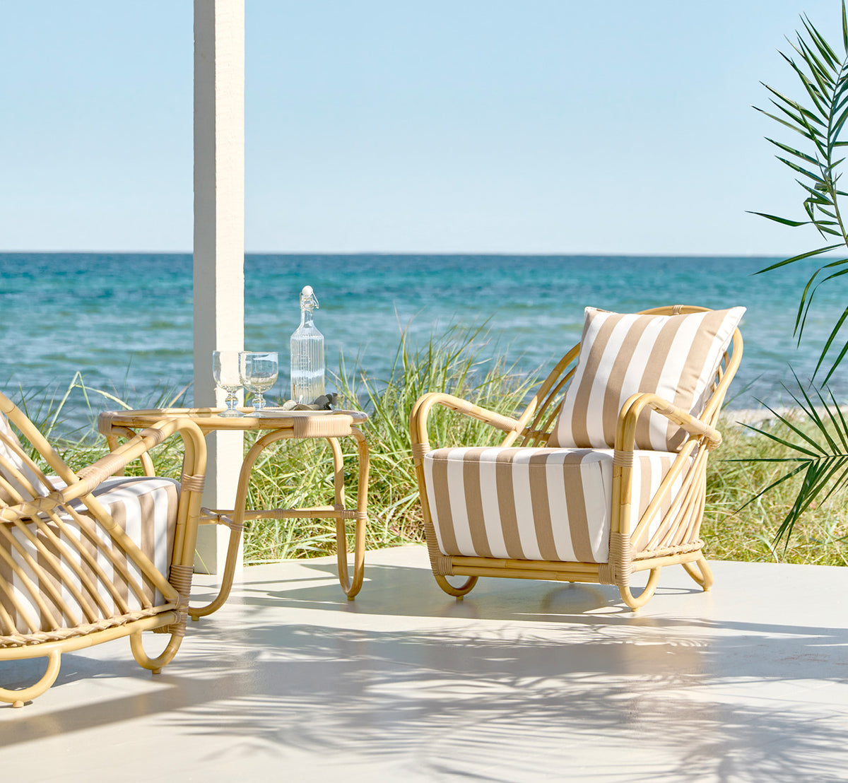 Dies ist ein Lifestyle-Bild von Charlottenborg Außen-Lounge-Sessel in natural Aluminium ArtFibre, auf einer Holzterrasse im Freien mit Blumen und.