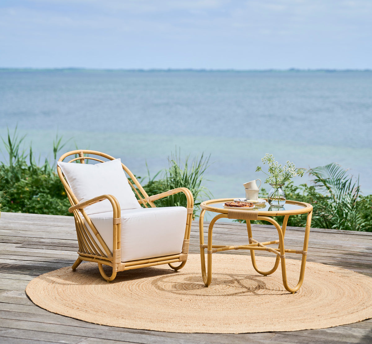 Dies ist ein Lifestyle-Bild von Charlottenborg Außen-Lounge-Sessel in natural Aluminium ArtFibre, auf einer Holzterrasse im Freien mit Blumen und.