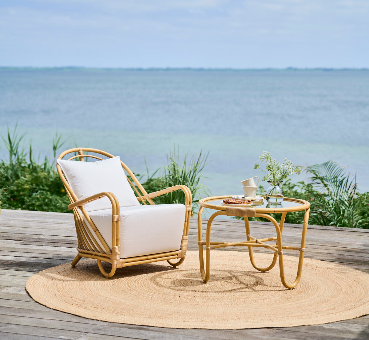 Dies ist ein Lifestyle-Bild von Charlottenborg Außen-Couchtisch in natural Aluminium ArtFibre, auf einer Holzterrasse im Freien mit Küstenbepflanzung.