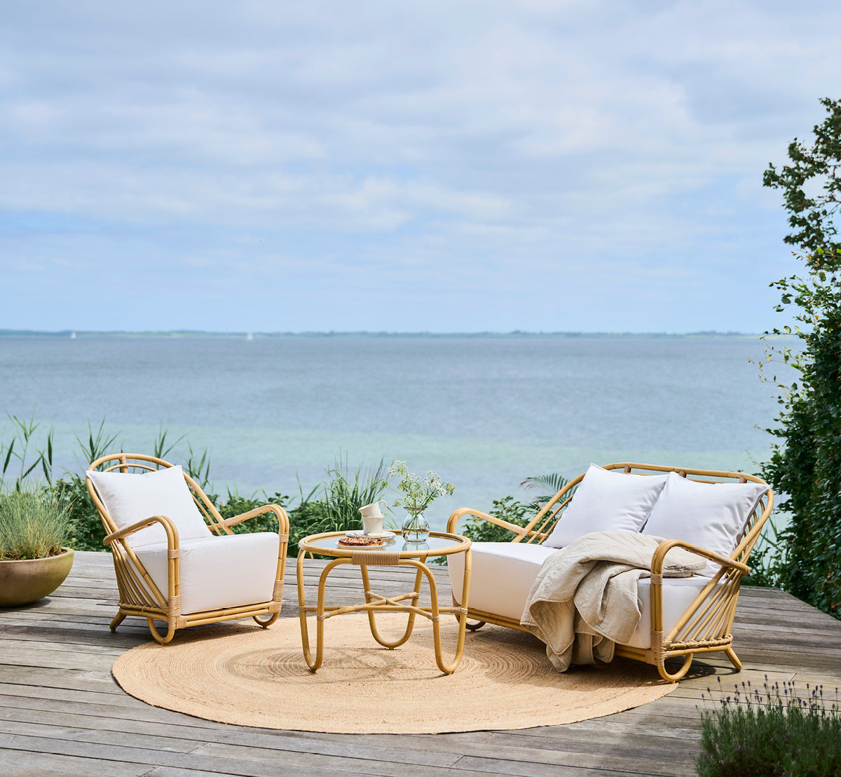 Dies ist ein Lifestyle-Bild von Charlottenborg Außen-Couchtisch in natural Aluminium ArtFibre, auf einer Holzterrasse im Freien mit Küstenbepflanzung.