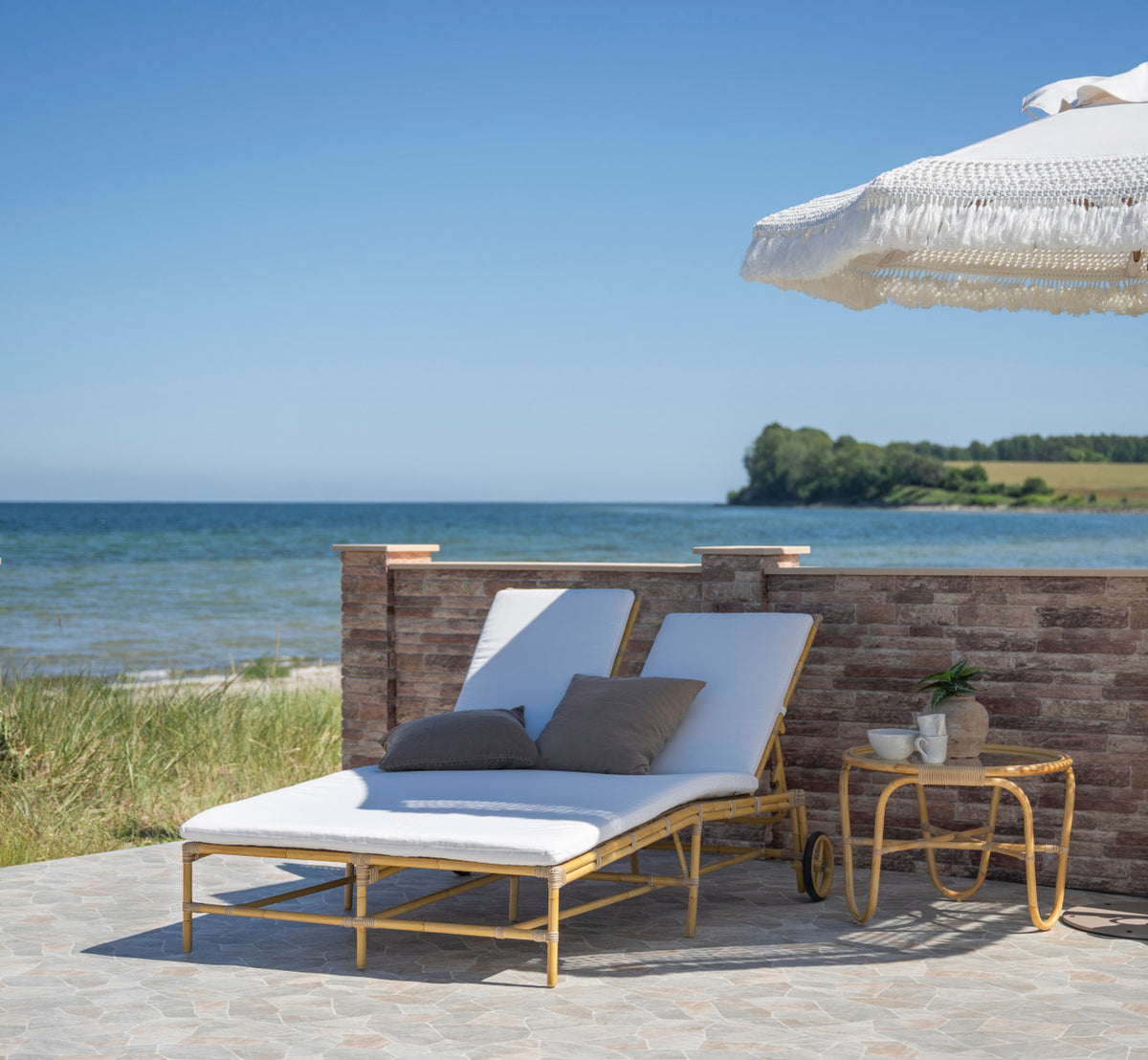 Dies ist ein Lifestyle-Bild von Charlottenborg Außen-Couchtisch in natural aluminium artfibre. Liege mit Kissen unter einem Sonnenschirm am Meer.