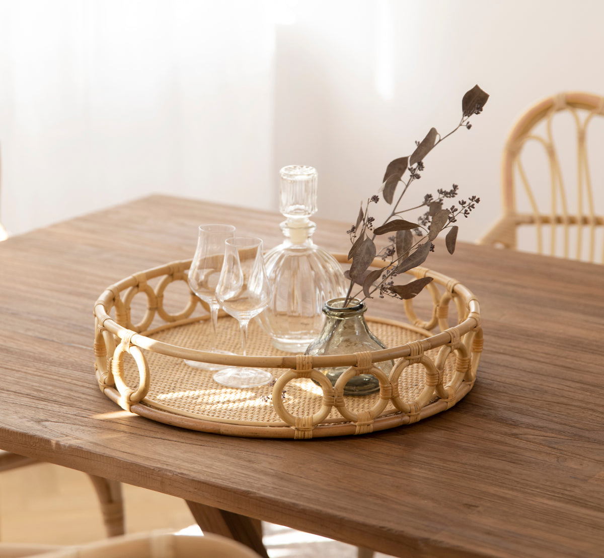 Dies ist ein Lifestyle-Bild von Mira Tray i natural rattan. Auf einem Teaktisch mit Kristallgläsern und einer Vase mit getrockneten Zweigen.