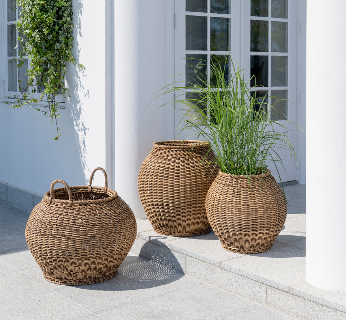 Dies ist ein Lifestyle-Bild von Smilla Korb in chestnut core weave ArtFibre Aluminium, im Freien auf einer sonnigen Terrasse mit weißer Mauer und.