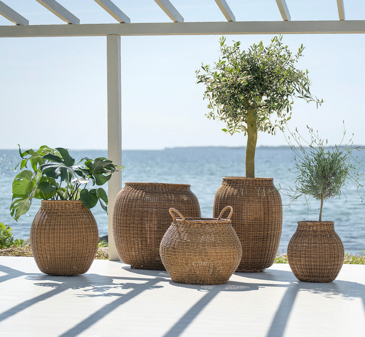 Dies ist ein Lifestyle-Bild von Alma Korb in chestnut core weave ArtFibre Aluminium, im Freien auf einer sonnigen Terrasse mit weißer Mauer und.