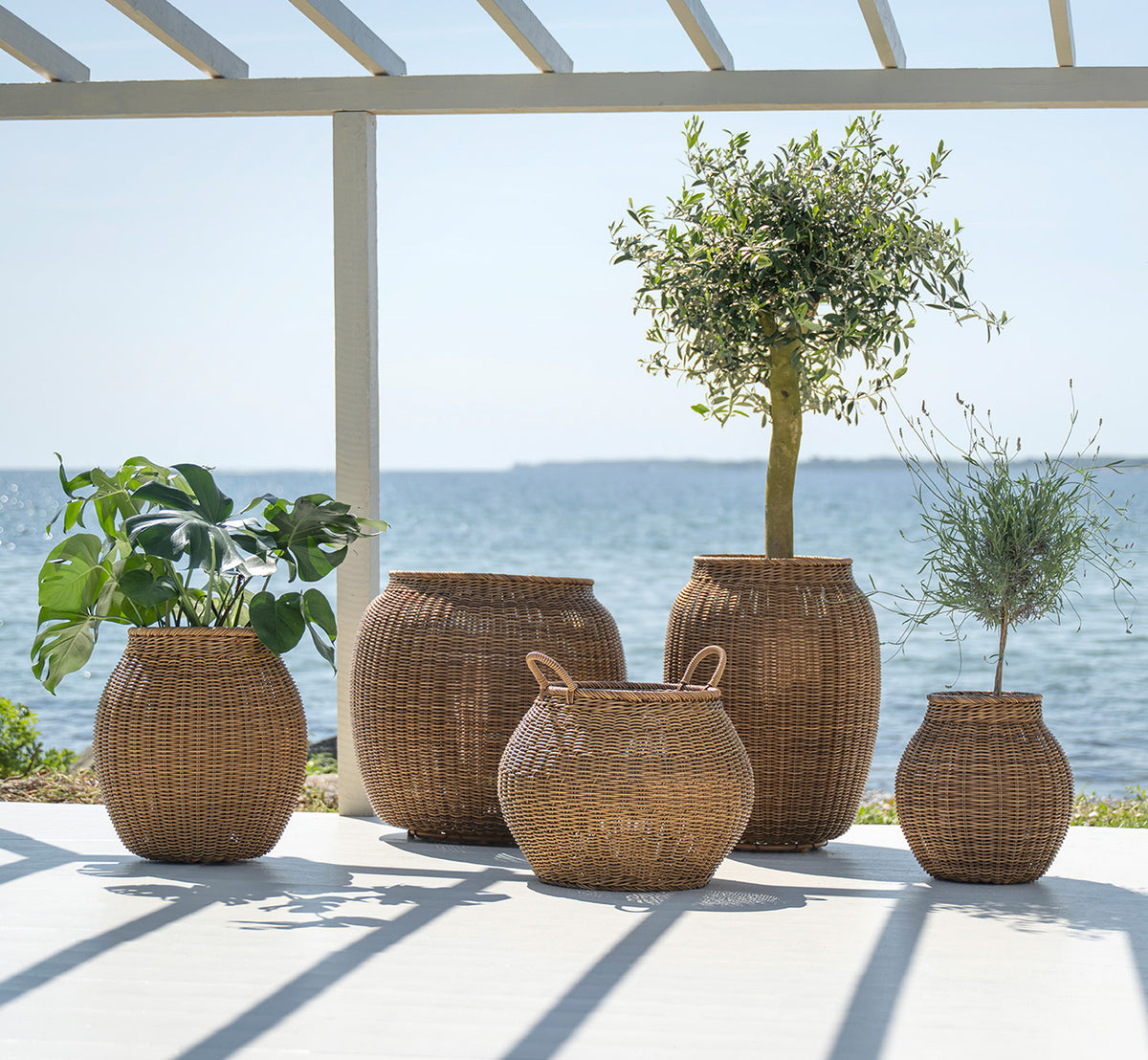 Dies ist ein Lifestyle-Bild von Alma Korb in chestnut core weave ArtFibre Aluminium, im Freien auf einer sonnigen Terrasse mit weißer Mauer und.