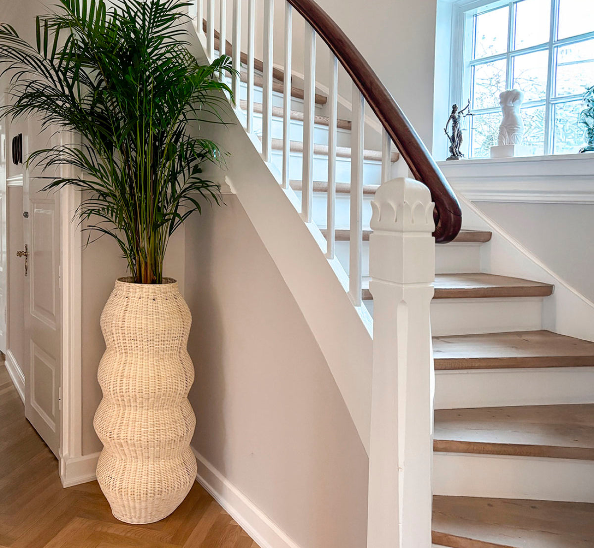 Dies ist ein Lifestyle-Bild von Unda Basket H102 cm in natural core weave iron frame. Als hoher Pflanztopf neben einer geschwungenen Treppe in einer hellen Halle verwendet.