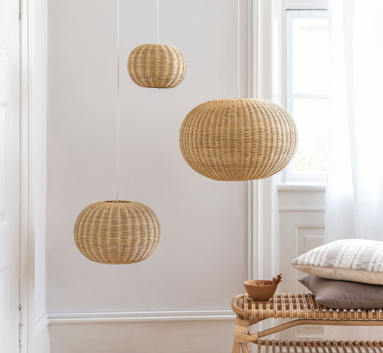 Dies ist ein Lifestyle-Bild von Tangelo Lampshade Ø49 cm | Medium in natural rattan. Drei Pendelleuchten vor einer weißen Wand über einer Rattanbank hängend.