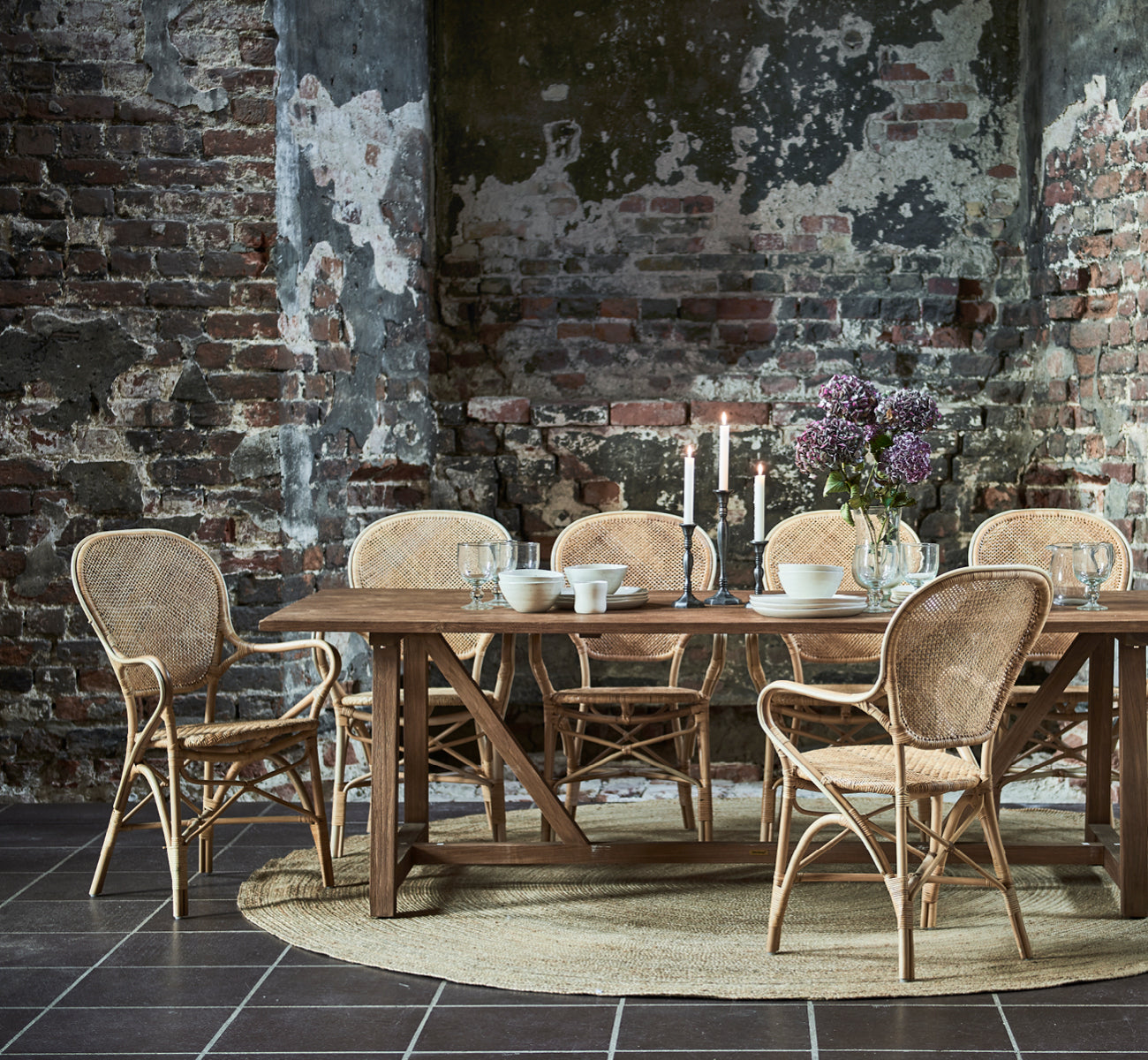 Dies ist ein Lifestyle-Bild von Jute Carpet Ø280 in natural jute. Unter einem Esstisch mit Rattanstühlen in einem rustikalen Raum mit sichtbarem Mauerwerk.