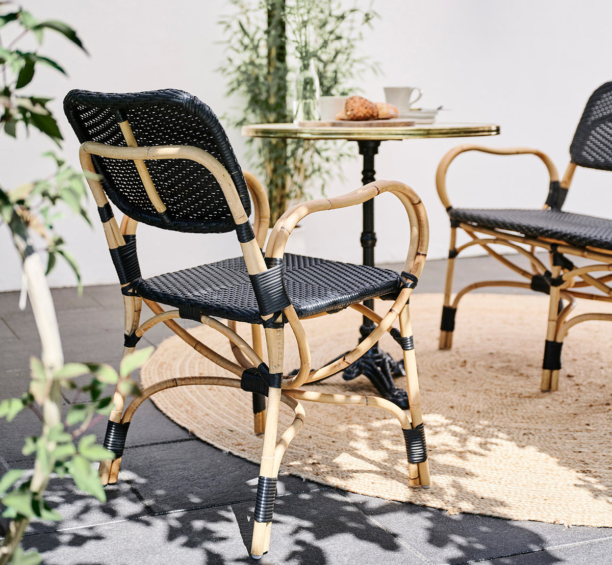 Dies ist ein Lifestyle-Bild von Bistro Dining Chair in skin on Rattan frame, in einem Außencafé-Ambiente mit einem Bistrotisch auf dunklem.