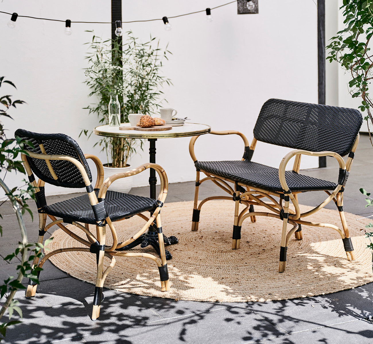 Dies ist ein Lifestyle-Bild von Bistro Bench in skin on Rattan frame, in einem Außencafé-Ambiente mit einem Bistrotisch auf dunklem Fliesenboden.