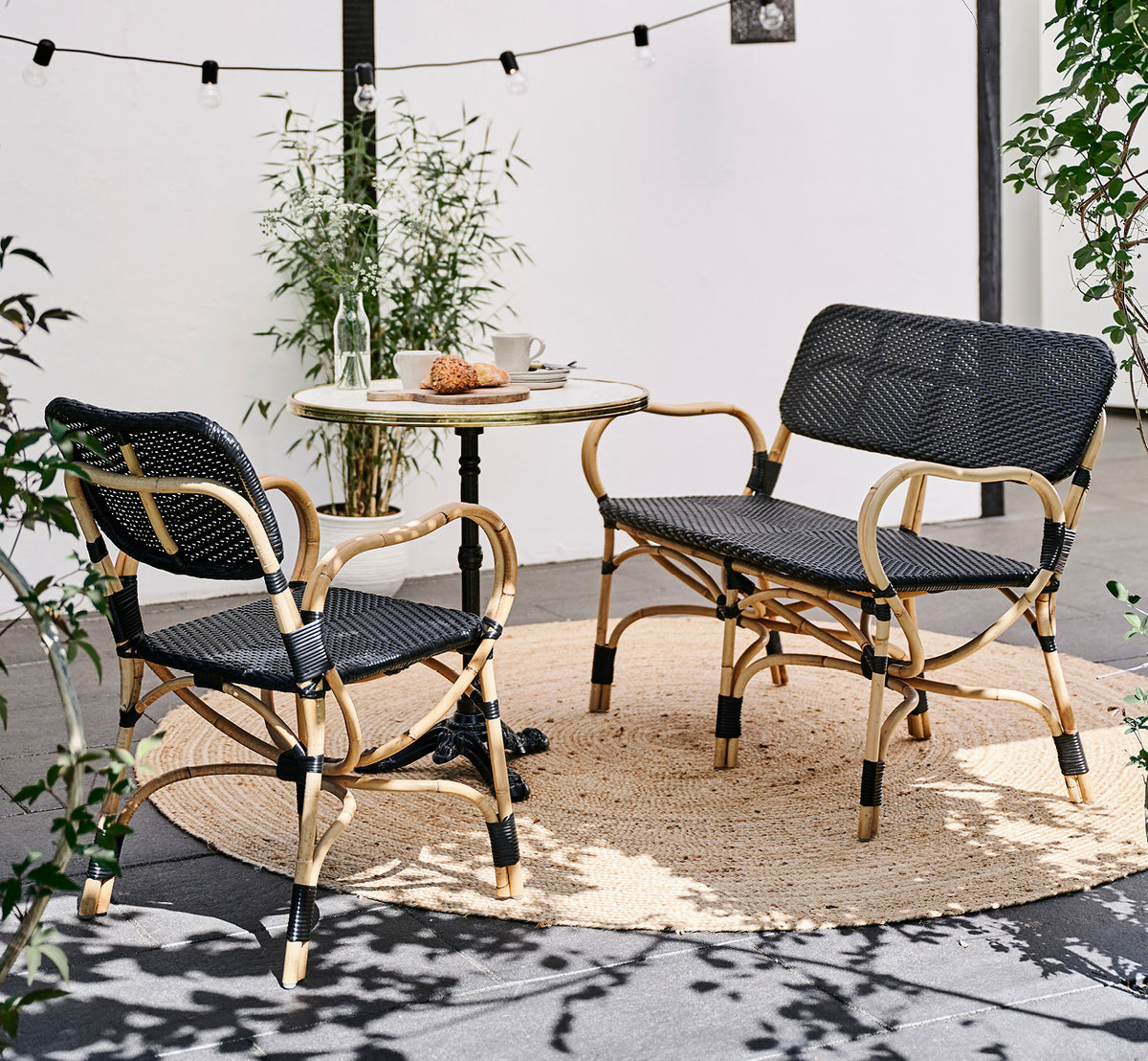 Dies ist ein Lifestyle-Bild von Bistro Bench in skin on Rattan frame, in einem Außencafé-Ambiente mit einem Bistrotisch auf dunklem Fliesenboden.