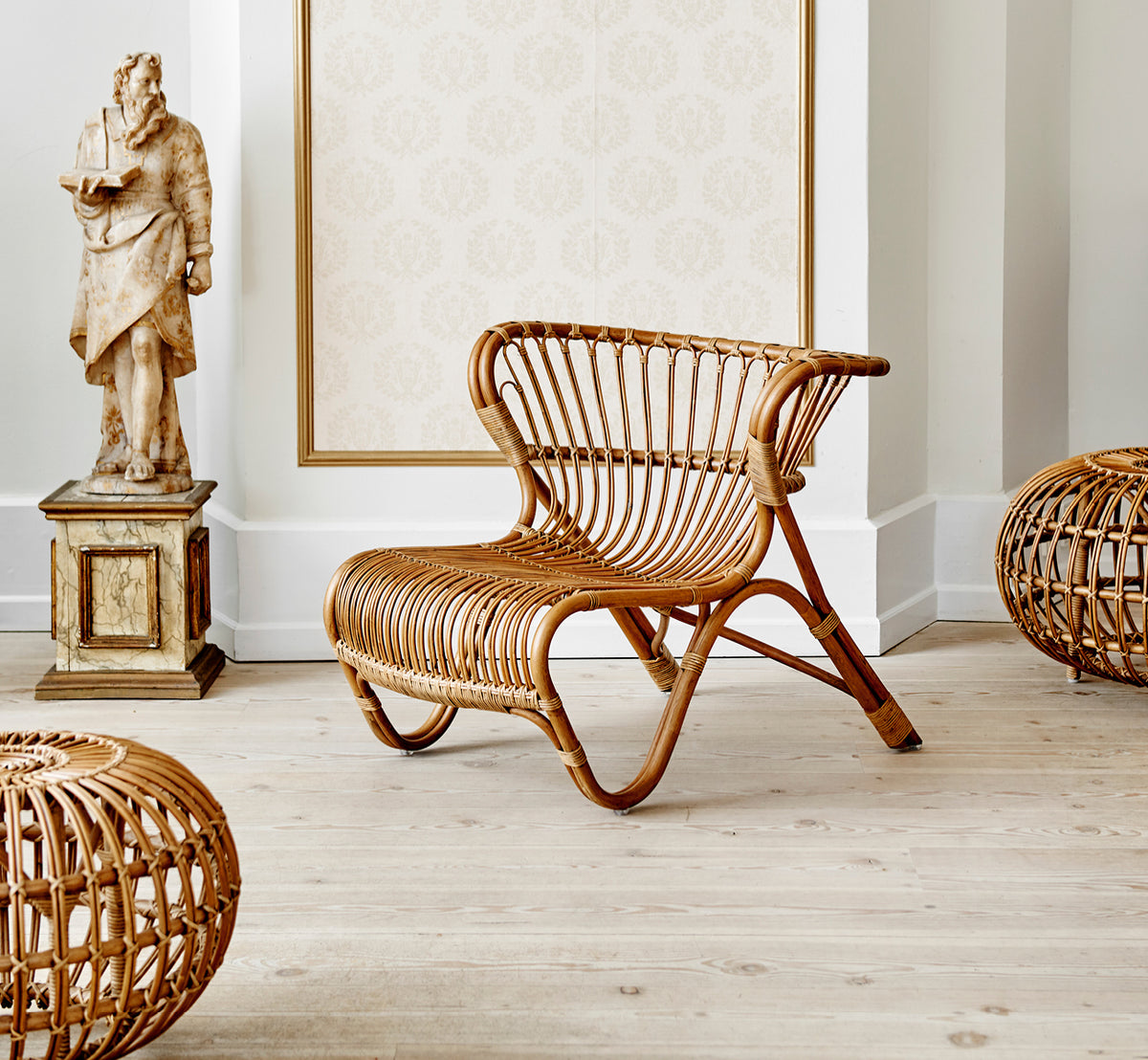 Dies ist ein Lifestyle-Bild von Ottoman Small in polished antique rattan. In einem Raum mit verzierter Tapete und einer klassischen Statue platziert.